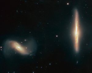 Two galaxies. The one at the right of the image is seen edge-on and vertical, glowing yellow-orange. The one at the left is a tilted disk, white and yellow near the center and reddish and white at the outsides.